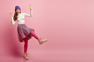 Funny girl dressed in long lush skirt, bright pink pantyhose and boots, foolishes around in studio during photoshoot, poses over pink background, wears fashionable attire, pretends falling down