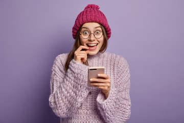 Communicative pleased female model holds cellphone, sends text messages, wears hat and sweater, isolated over purple background, connected to wireless internet, enjoys free time and nice winter