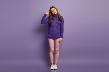 Positive young female foolishes indoor, imitates gun shoot, keeps hand near temple, tilts head and smiles broadly, wears purple loose sweater, trousers and sneakers. Full length shot. Body language