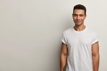 Smiling young Caucasian man with pleased expression, dressed in casual clothes, has muscular body,...