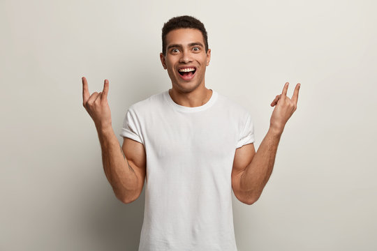 Happy pleased man with cheerful facial expression, makes rock n roll gesture, enjoys cool music, dressed in white t shirt, poses in studio, makes horns, has short dark hair. Body language concept