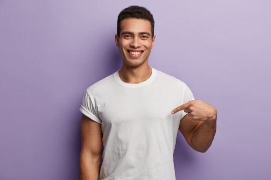 Look At My New T Shirt. Indoor Pleased Caucasian Man With Short Haitcut, Points At Blank Space Of His Clothes, Smiles Positively, Isolated Over Purple Background. Advertisement And Clothing Concept