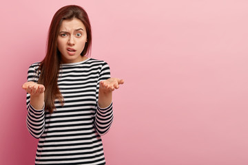 Indoor shot of young indignant woman gestures with indignation, frown face, wears striped sweater,...