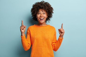 Horizontal shot of positive optimisitc African American woman points with both fore fingers upwards, wears casual jumper, advertises new item, isolated over blue background. Look at this thing