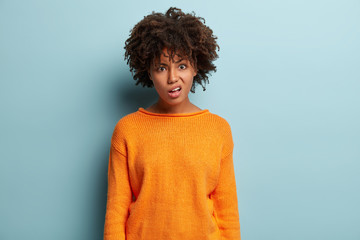 Displeased woman with dark skin, Afro hairstyle, purses lips, feels discontet, looks with indignation, wears orange oversized jumper, isolated over blue background. Negative emotions concept