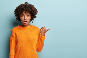 Studio shot of astonished black mixed race woman points with thumb aside, wears orange jumper, keeps jaw dropped, has surprised facial expression, isolated over blue background. What is there I wonder
