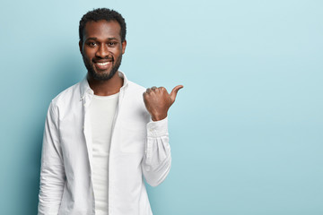 Photo of handsome guy has brilliant smile, shows white perfect teeth, wears stylish clothing, attracts your attention away, points with thumb, isolated over blue background. Use this copy space