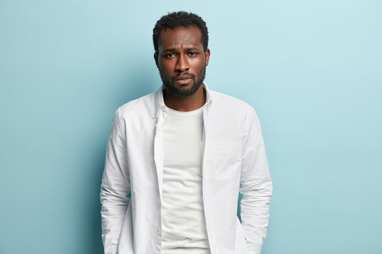 Image of sad sorrowful black man with thick bristle, wears white shirt, feels troublesome, looks at camera with miserable expression, isolated over blue background. People, melancholy, problem concept