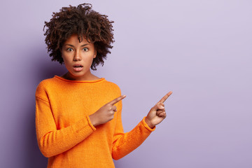 Shot of puzzled dark skinned girl in casual jumper, points with both fore fingers at empty space,...