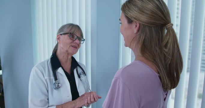 Over the shoulder shot of doctor giving news to female patient by window. Senior primary care physician speaking with Caucasian woman in hospital. Slow motion 4k