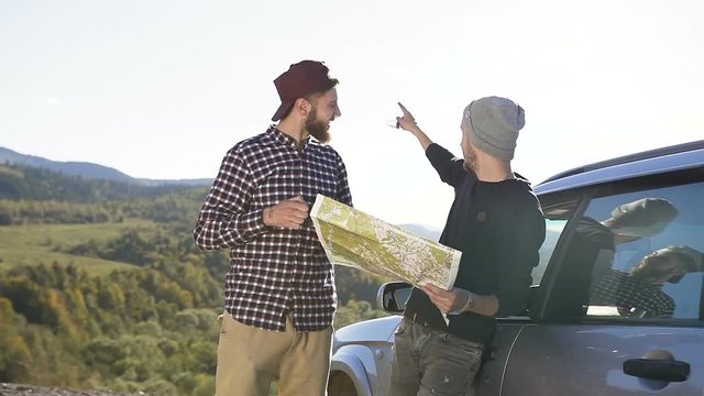 Two friends guys standing near the car drinking tea while looking at the roadmap making a further route to climb the mountains, laughing and talking. Travel in the mountains by car. Nature, outdoors