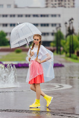 Young pretty girl with two braids in yellow boots and with transparent umbrella stands near fountain.