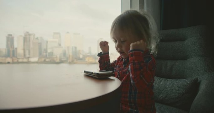 Little toddler playing a game on a smartphone by window in city