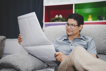 Asian senior man relaxing and reading Business News newspaper on sofa in living room at home