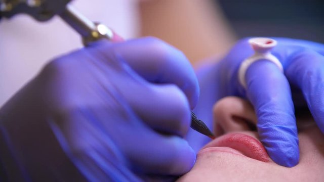 Lips tattoo. Cosmetologist applying permanent makeup tattoo on lips. Professional tattooist making permanent make up. Close-up