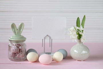 Easter holiday.Spring  Easter Mockup.decorative jar with bunny ears easter eggs, white board, hyacinth flower on a pink table on a white wooden background.Easter mood. 