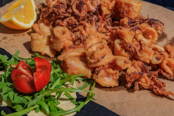 Crispy fried calamari rings and tentacles served on a rustic board on a piece of paper with lemon wedge and a salad of arugula and tomato with olive oil at a restaurant in Ventimiglia, Italy