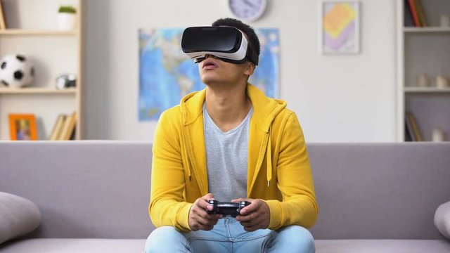 Afro-American teenage boy playing video game in VR headset, modern technologies