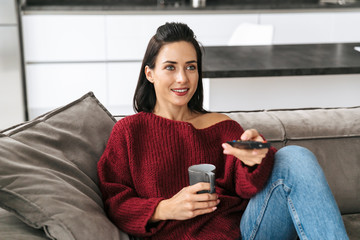 Woman indoors in home on sofa watch TV drinking coffee.