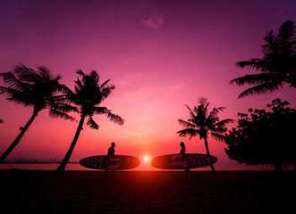 Silhouette of surfers couple holding long surf boards at sunset on tropical beach