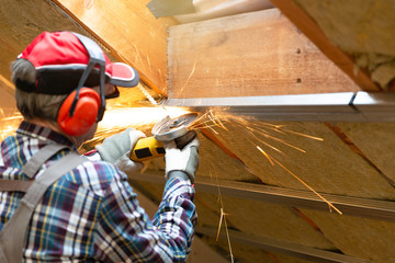 Man fixing metal frame using angle grinder on attic ceiling covered with rock wool