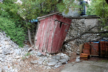 Destroyed cracked traditional old outdoor wooden toilet leaning to one side with broken dilapidated boards surrounded with rocks and construction material with trees in background on warm sunny day
