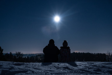 Silhouette of a man and woman sitting on a snow covered hill at night and looking at the moon