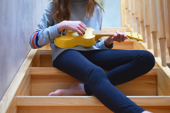 Bare foot teen girl wearing black leggings and grey shirt playing song on yellow ukulele guitar while sitting on wooden stairs steps at home indoors. Music and hobbies concept.