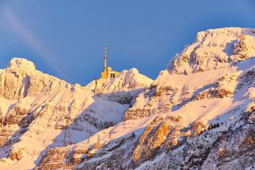 Säntis im Winter, Bergstation mit Sendemast, schneebedecktes Felsmassiv im gelben Abendlicht, blauer Himmel, Seilbahnmasten