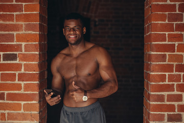 Fototapeta na wymiar African shirtless sportsman holding his smartphone, standing in brick wall gym entrance and looking at camera before starting workout. Modern technologies, healthy lifestyle concept, copy space
