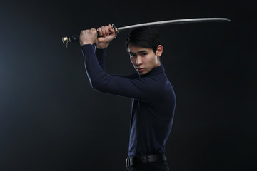 Obraz na płótnie Canvas A young Asian man with a katana in his hands against a dark background.