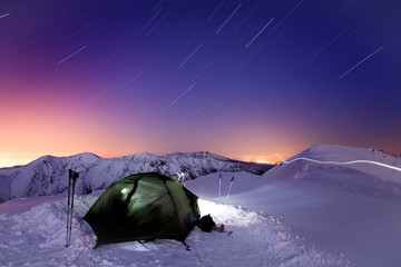 Winter camp, night, shining green tent in the snow. Night shot, long exposure, sleeping in the snow outside. Alps mountains landscape panoramic view. Wonderful winter day in Switzerland, Europe. 