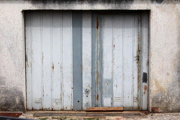 Old dilapidated partially rusted grey metal garage doors mounted on concrete wall next to concrete road on warm sunny day
