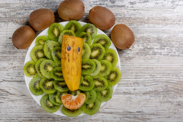 The face of fruit, kiwi, banana, a slice of mandarin on a white plate. Wooden light background, Flat Lay, Copy space.