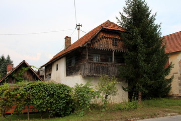 Old stone and wood family house with wooden balcony in front next to tall pine tree surrounded with uncut grass and garden vegetation on warm sunny day