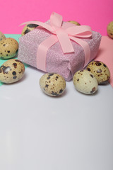 Obraz na płótnie Canvas Quail eggs lie on the white surface. Next to the box with a gift wrapped in wrapping paper and tied with a ribbon. Sheets of colored paper for background.