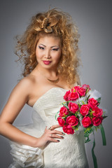 young girl Asian looks-bride in white dress with a bouquet of flowers shows a beautiful wedding hairstyle on a gray background