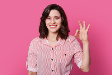 Photo of optimistic joyful beautiful woman has professional makeup, wavy dark hair, makes okay gesture, wears striped shirt, isolated over pink background. Body language concept. People and style