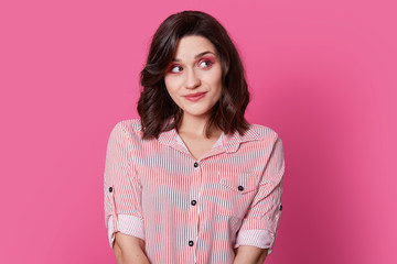 Fototapeta premium Dreamy pleased lady with short hair, looks aside with thoughtful expression, contemplates about something, dressed in fashionable attire, models over pink background. Retro woman models indoor