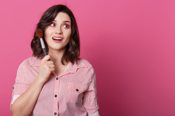 Fototapeta premium Attractive female model keeps gaze aside, has pink makeup, curly hair, holds cosmetic brush, works in beauty salon, wears striped shirt, isolated over rosy background with blank space aside.