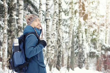 Handsome bearded male hiker in warm clothes with backpack and thermos travel in winter snowy forest. Seasonal outdoor travel concept. Copy space