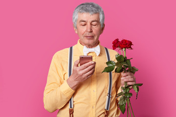 Isolated shot of attractive old man focused in smartphone, recives message from lover, agree to have date, carries red roses, wears yellow shirt, suspenders and bowtie, isolated over pink background
