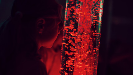 Child in therapy sensory stimulating room, snoezelen. Child interacting with colored lights bubble tube lamp during therapy session. - obrazy, fototapety, plakaty