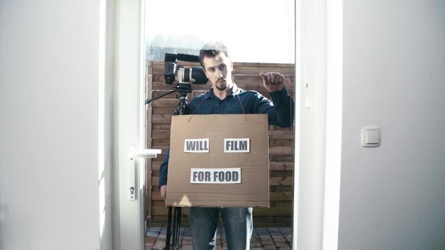 Man hold meme sign Will Film For Food 4K. Static shot of a male person in front of a glass door knock and holding a sign with words.