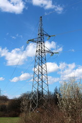 Tall metal electrical power line utility pole made of strong metal pipes with multiple electrical wires connected with glass insulators soaring above trees with cloudy blue sky in background