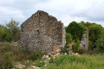 Traditional stone house ruins with missing roof partially overgrown with crawler plant surrounded with dense vegetation on warm cloudy day