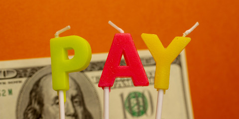 Horisontal Banner of Concept word Pay from festive multi-colored wax paraffin candles with dollar bill on orange background.