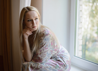 Pensive girl in bathrobe sitting on the windowsill. Natural light.