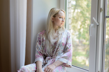 Pensive girl in bathrobe sitting on the windowsill. Natural light.