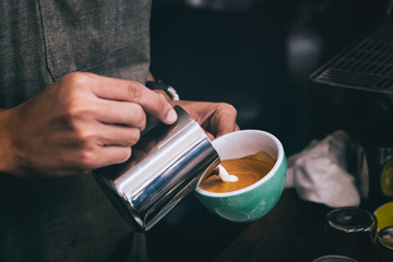 Close-up of male barista hand holding and pouring hot milk for prepare latte art on a green cup of cappuccino coffee. vintage color tone.